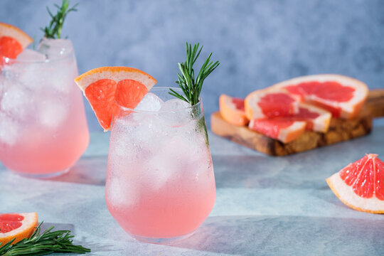Two glasses filled with pink drinks containing grapefruit slices and ice sit on gray background. In the background, there are more grapefruit slices on a wooden board. 