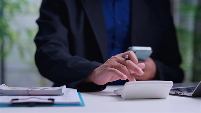 Male entrepreneur calculating financial budget holding mobile phone planning investment sitting beside paper currency stack laptop clipboard office desk workspace environment.