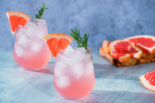 Refreshing pink grapefruit mocktail served over ice and garnished with a fresh grapefruit wedge and rosemary sprig. Two elegant glasses of sparkling citrus beverage on a blue textured background.