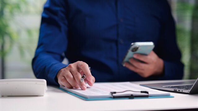 Businessman holding pen over clipboard analyzing financial spreadsheet papers, holding smartphone, sitting desk office workspace, managing corporate budget, calculating revenue, checking tax data.