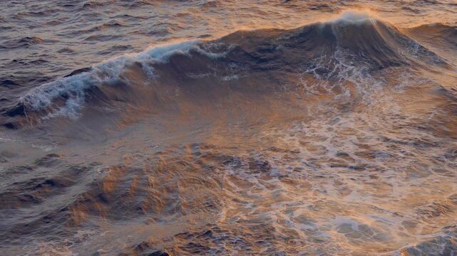 Caribbean Sea waves roll gently through golden sunset light as warm orange and amber tones illuminate the textured water surface. White foam crests form and dissolve across the deep blue ocean.