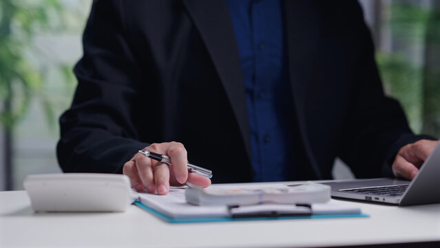 Male accountant analyzing financial document holding pen near laptop calculator money stack inside office workplace desk calculating business tax cost planning investment profit.