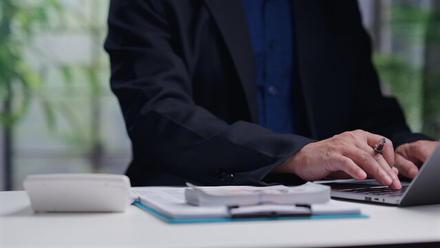 Professional accountant types laptop keyboard, analyzes financial report document, calculates business tax, manages cash money budget, works indoor office desk, holding metal pen.