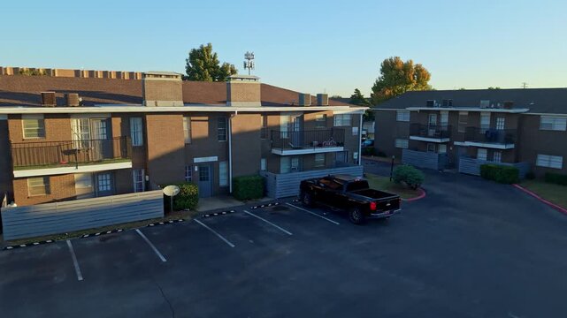 Evening aerial view of a brick multifamily housing complex with satellite dishes, parked cars, and fenced areas.
