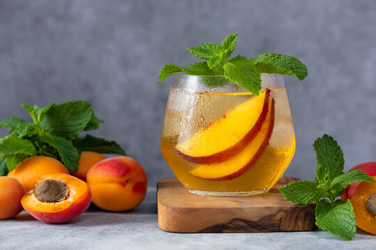 Close-up refreshing summer apricot and nectarine cocktail served over ice with fresh mint leaves in a modern glass. The drink is garnished with juicy stone fruit slices and surrounded by ripe apricots
