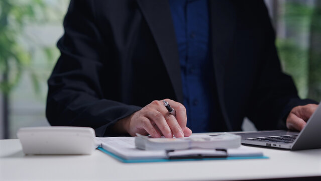 Corporate accountant wearing black jacket calculates business budget, holding pen, typing laptop keyboard, analyzing paper document stack, managing cash money inside office workspace.