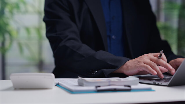 Businessman wearing dark suit operates laptop keyboard near paper document, money stack, calculator inside office workspace, analyzing financial data, calculating budget, planning investment.