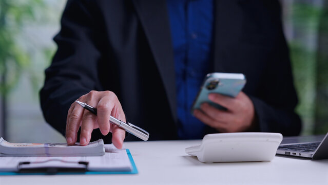 Male entrepreneur wearing dark blazer holds silver pen over cash money stack near calculator, checking financial ledger document, holding mobile phone inside office workspace.
