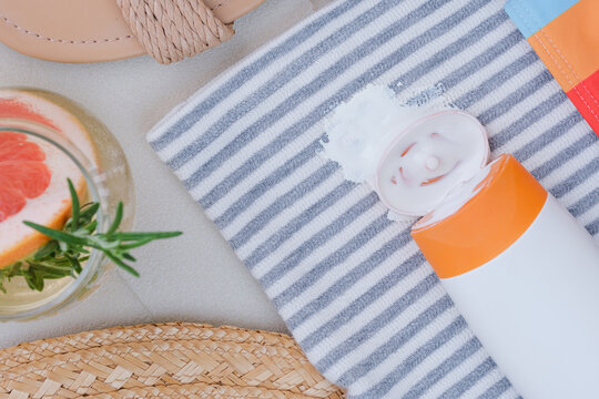 Sunscreen spilled on a towel with stripes. A drink with a slice of grapefruit and herbs sits nearby. Sandals are visible on the side, showing a relaxed beach scene. top view