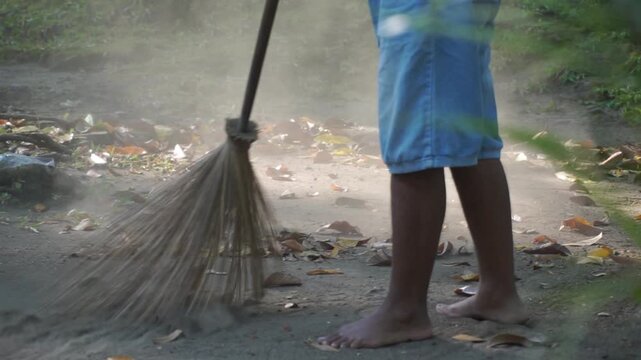 Slow motion of Action shot of a person sweeping dry leaves on dirt ground with a traditional broom stick, showing flying dust particles, motion blur, and outdoor yard cleaning. Office boy