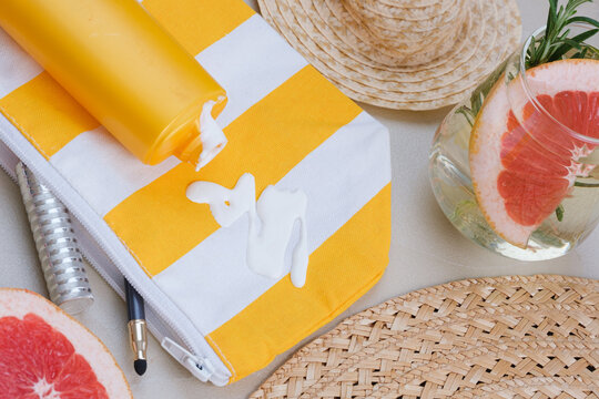 An open cosmetic bag with spilled sunscreen is surrounded by a straw hat, beach sandals, half a grapefruit, a makeup brush, and cosmetic accessories on the sand. top view