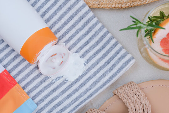 Sunscreen spilled on a towel with stripes. A drink with a slice of grapefruit and herbs sits nearby. Sandals are visible on the side, showing a relaxed beach scene. top view