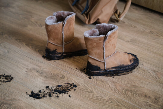 A pair of tan suede winter boots covered in wet black mud standing on a wooden floor inside a home, leaving dirty smudges and scattered clumps of soil. Home cleaning, family life, and everyday messes 