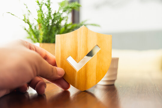 Real estate insurance and property protection concept. Hand holding wooden shield with check mark in modern home. Ideal for house mortgage, rent guarantee, and secure investment business.