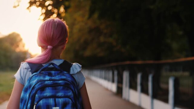 Young girl with pink hair walks down a tree-lined path carrying a blue backpack at sunset, cinematic footage.