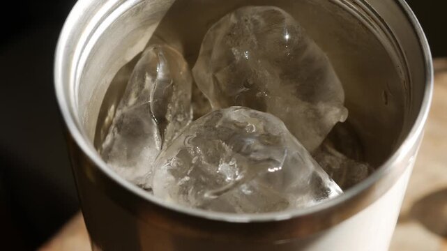 Close-up top angle view into stainless steel tumbler cup with melting ice cubes in golden warm light on wooden table in cafe
