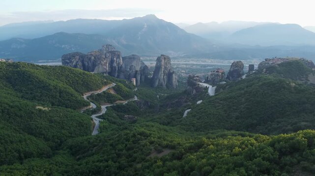 Drone aerial footage of the Meteora monasteries and towering sandstone rock formations overlooking Kalabaka, Greece, captured during golden hour with panoramic mountain scenery.
