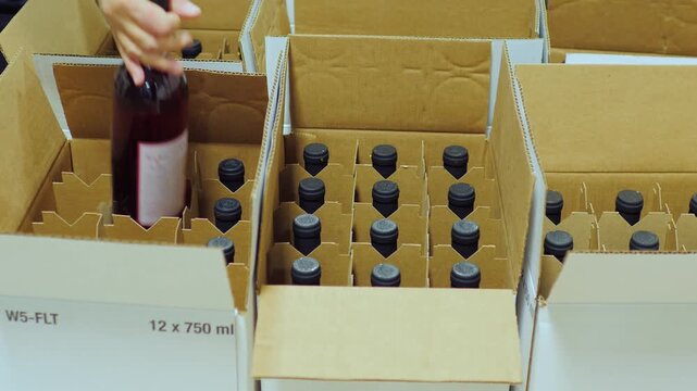Hands packing wine bottles into cardboard boxes at warehouse, preparation for shipping.