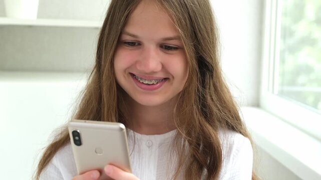 Happy beautiful teen girl with dental braces on teeth holding mobile phone texting using mobile app. Smiling teenager in brackets using cellphone