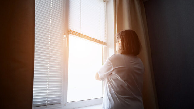 Woman standing by a window with blinds, looking at bright sunlight from outside, contemplating thoughts of future and opportunities with a sense of hope and new beginnings