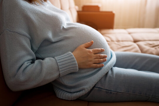 Expectant mother in soft home clothes lies on a couch, gently holding her belly. Warm natural light and a calm, comfortable living room create a peaceful maternity mood