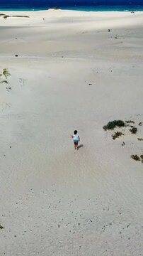 High angle drone view of a fit woman running barefoot on the soft white sand of a beautiful deserted beach, enjoying a sunny summer day while on vacation near the turquoise ocean waves