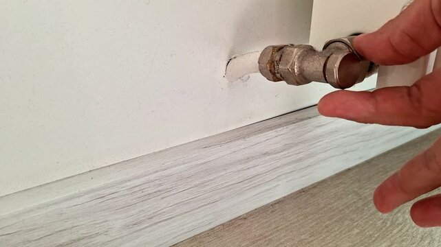 A hand carefully unscrews a radiator valve connection near the wall baseboard during a home plumbing maintenance task. Ideal footage for heating systems, repairs, renovation, and DIY projects