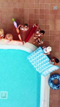 Aerial view of a group of attractive young women in bikinis having fun together with inflatable mattresses and floats, celebrating a summer party by the pool, waving hands and smiling