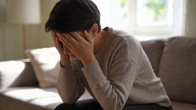 A woman sits on a couch in a living room, holding her head in her hands, feeling distressed.