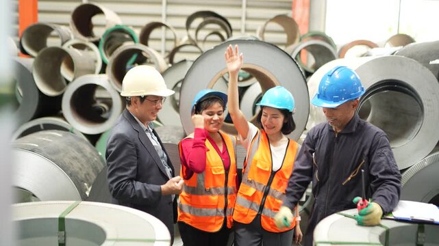 Engineering team celebrating successful production milestone, hands together beside steel coil inventory in industrial warehouse. Manufacturing teamwork, workplace collaboration and business success.