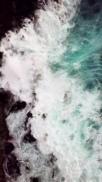 Beautiful turquoise ocean waves breaking against a dramatic dark rocky coastline, creating white foam and spray from a stunning top-down aerial perspective showing the power of nature