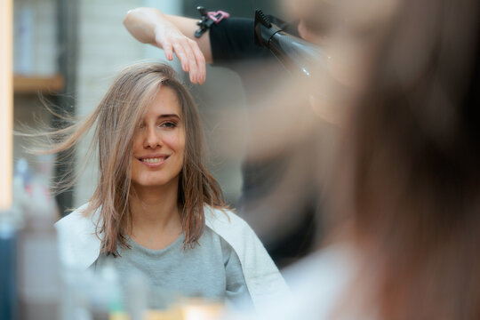 Professional Hairdresser Drying Beautiful Young Woman Hair With Blow Dryer. Smiling client enjoying a professional blow dry styling treatment from her hairstylist at beauty salon
