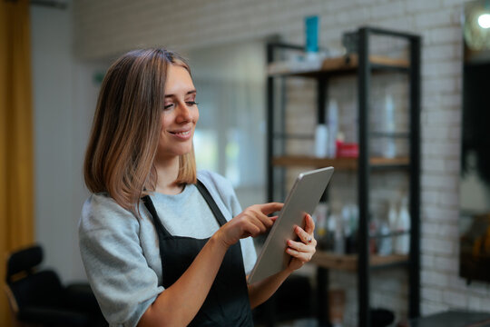 Smiling Hairdresser In Black Apron Checking Appointments On Digital Tablet. Happy hairstylist holding a tablet computer while standing inside a modern beauty salon workstation