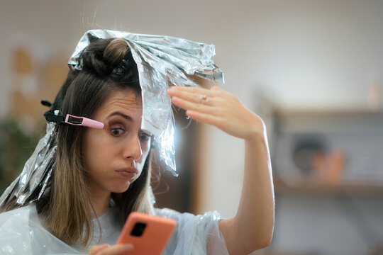 Stressed Woman With Hair Foils Reading Text Message At Salon. Concentrated female customer looking at smartphone messages while getting a balayage hair dye treatment 