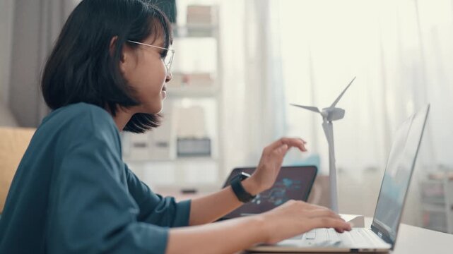 A student types on a laptop in a bright room. The student focuses on schoolwork while a small wind turbine model sits nearby. Natural light fills the space, creating a workspace.