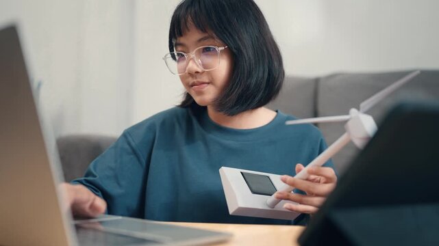 A student types on a laptop in a bright room. The student focuses on schoolwork while a small wind turbine model sits nearby. Natural light fills the space, creating a workspace.