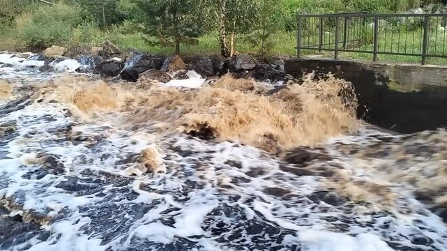 Rushing muddy floodwater flowing rapidly past concrete dam spillway after heavy rain storm