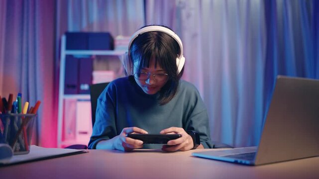 A girl sits at a desk in her room during the evening. She focuses on her mobile game while wearing headphones. A laptop and stationery are on the desk.