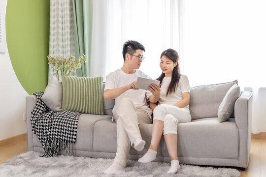 A young Asian couple sits on a sofa in a bright living room, jointly viewing a tablet and interacting casually.