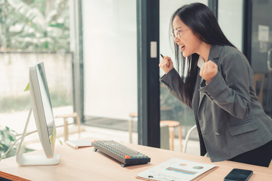 Excited Asian businesswoman celebrating success raising fists while looking at computer screen.