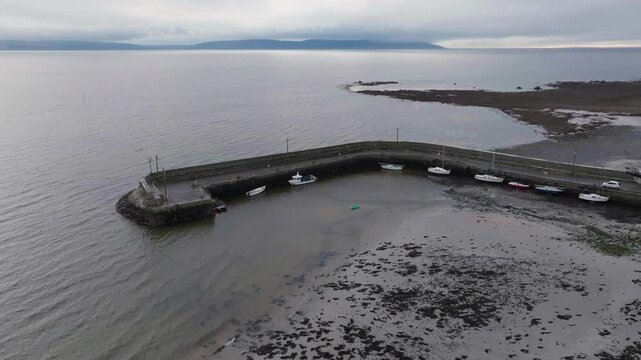 A cinematic aerial view of the historic Barna stone pier in Ireland, featuring small fishing boats at low tide with the vast Galway Bay and distant mountains under a cloudy sky.