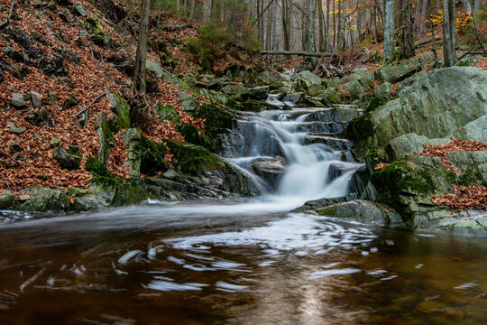 Small waterfall cascading over mossy rocks in a peaceful autumn forest with fallen orange leaves