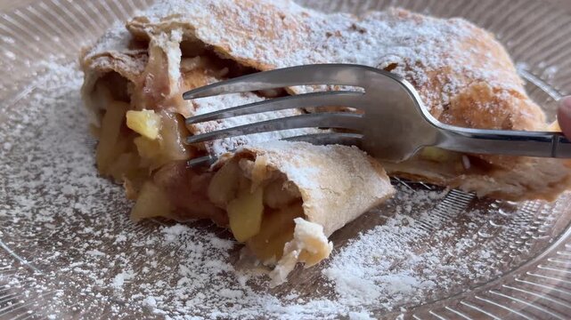 Fork cutting homemade apple strudel with juicy apple filling and powdered sugar, close up dessert serving