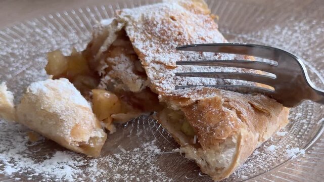 Fork breaking homemade apple strudel with juicy apple filling and powdered sugar, close up dessert