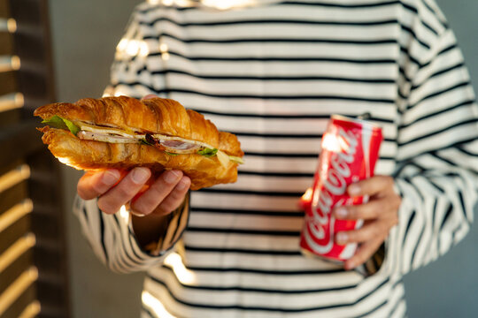 Minsk, Belarus - July 30, 2026: Horizontal shot of a person pouring Coca-Cola from a red aluminum can into a tall glass with a striped straw next to a croissant sandwich