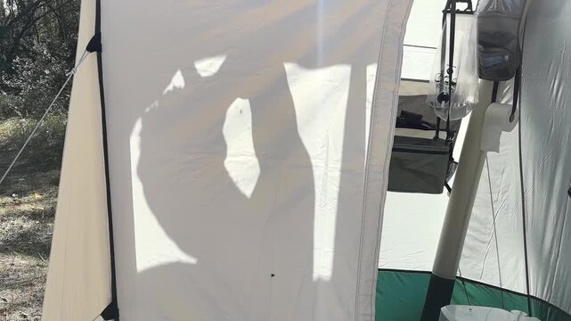 A man is silhouetted behind a tent as he washes himself at a peaceful campsite. The sunlight creates beautiful shadows, enhancing the tranquil atmosphere of the outdoors.