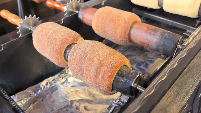 In a vibrant street market, the sweet aroma of freshly baked trdelnik fills the air. Cooks expertly roll dough onto rotating posts, creating a delicious treat.