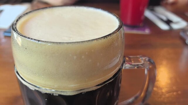 Foamy dark beer sits invitingly in a classic mug as friends gather in a warmly lit pub. The atmosphere buzzes with chatter, laughter, and the clinking of glasses.