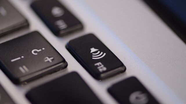 Extreme close up shot of a finger pressing the volume up button on a silver laptop keyboard. Representing increasing sound volume, listening to music. Pushing the volume button.
