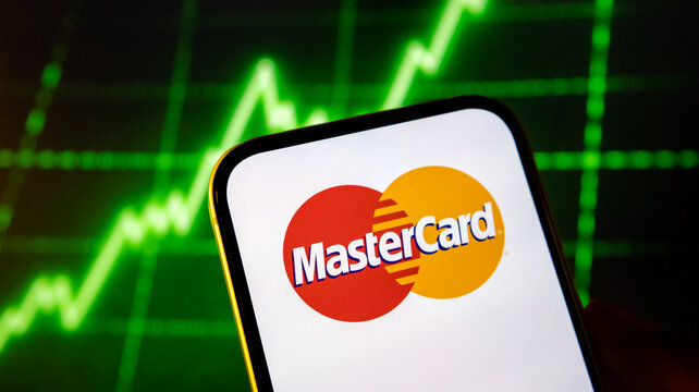 Mastercard logo on phone screen with financial stock market graph background.